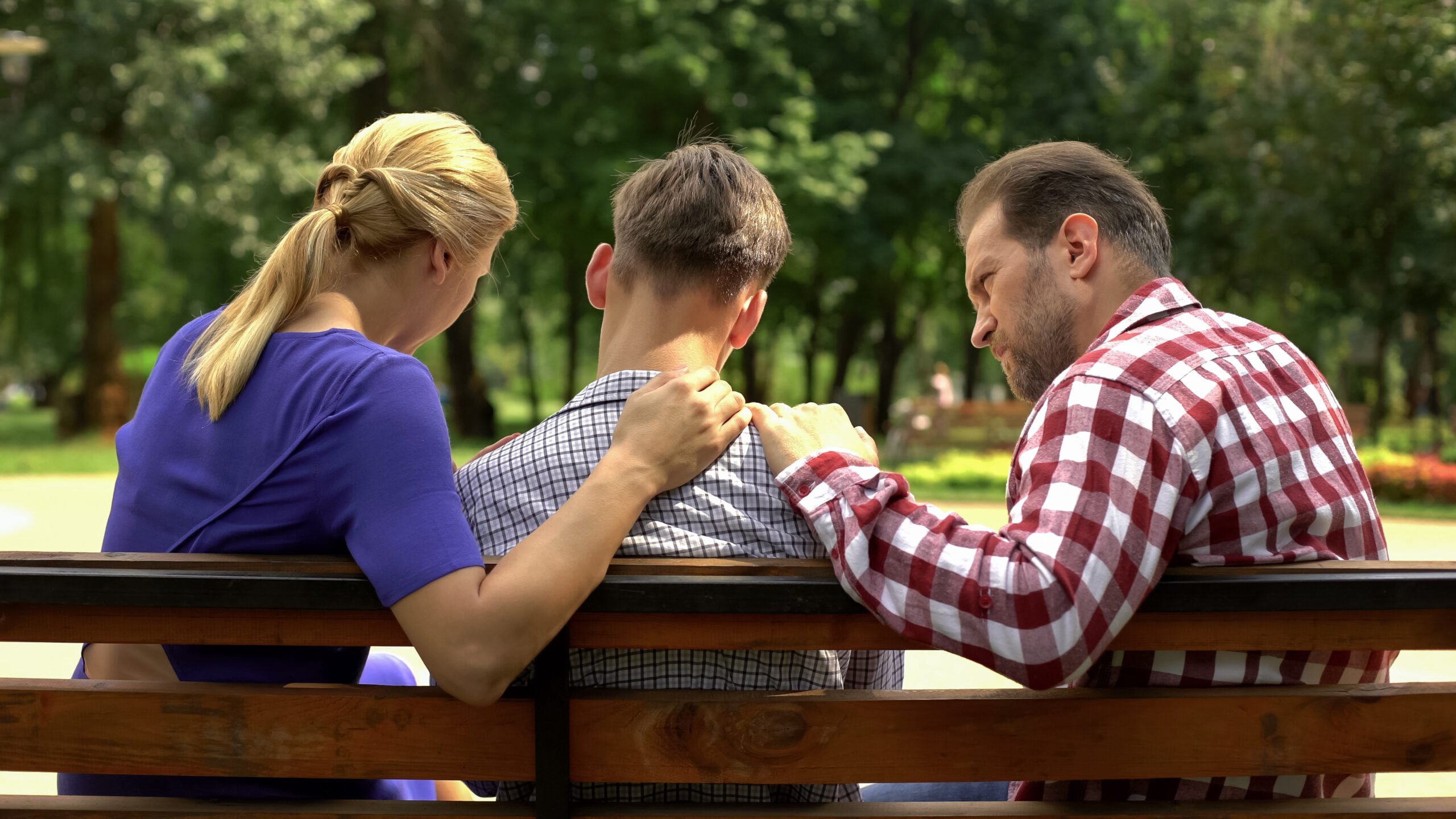 Caring mother and dad supporting sad teen son sitting on bench in park, consoling after the funeral