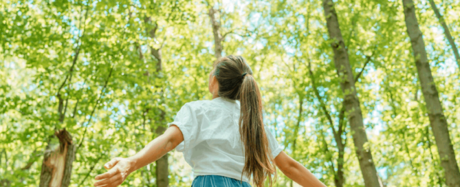A woman standing in a sunlit forest with her arms outstretched, surrounded by tall green trees.